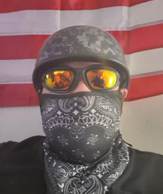 Person wearing a low-profile carbon fiber beanie half helmet from beaniehelmets.com and riding goggles with a black shirt and a bandana over their mouth against an American flag background.