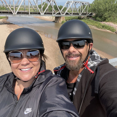 Two people on a road trip wearing matte black polo-style beanie helmets from beaniehelmets.com and sunglasses, standing by a river with a bridge in the background.