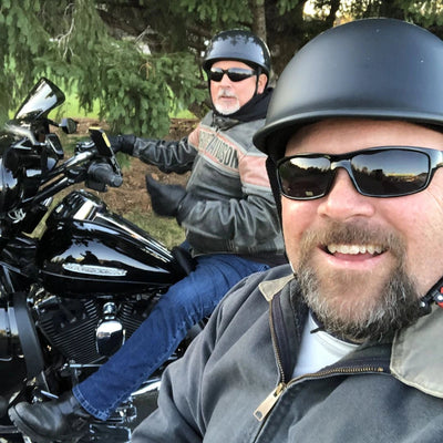 Two men on motorcycles wearing polo style beanie helmets with trees in the background