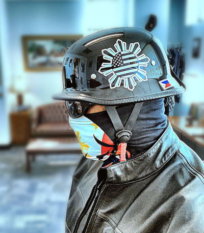 A biker wearing a black Mayan-style Germanator helmet with USA flag, sunglasses, colourful face mask, and a black leather jacket. Indoor settings.
