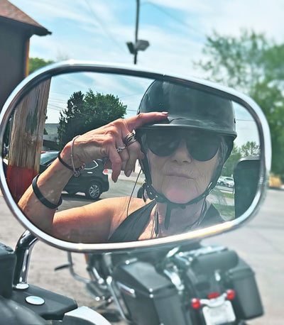 Lady biker wearing a gloss black polo-style beanie helmet and sunglasses reflected in a motorcycle side mirror.